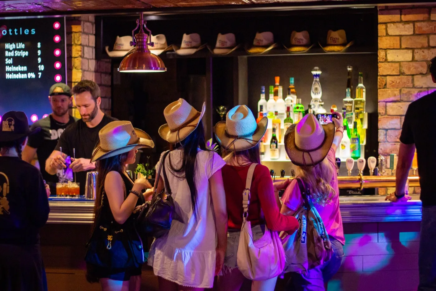 Cowgirls in straw hats ordering drinks at the bar as the bartender mixes a cocktail